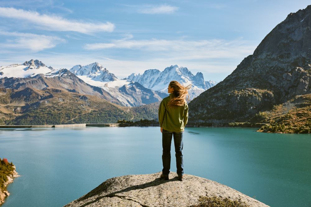 Woman on rock staring at mountains and lake A woman is standing on a rock with a big blue mountain lake beneth hair. She is looking to a vast mountain landscape