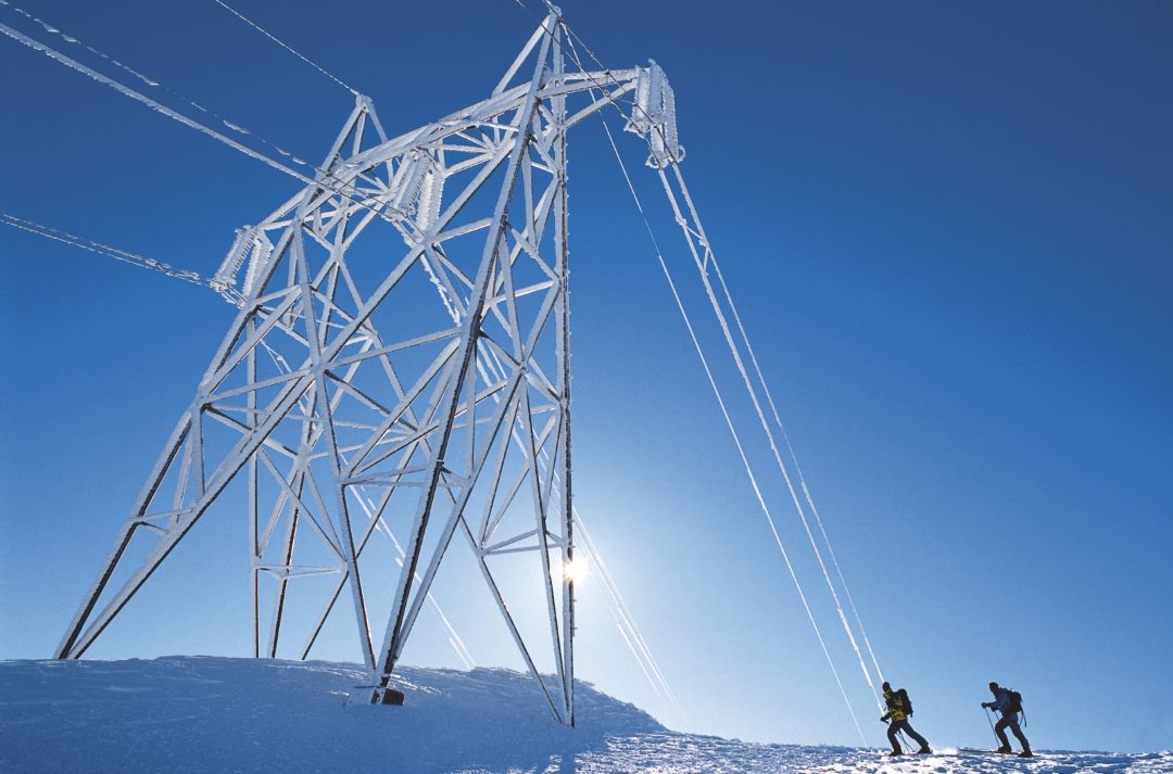 A big electricity pole is coverd in snow while to hikers are to its right