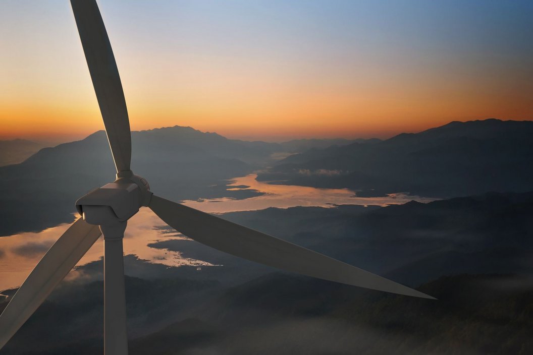 There is a big windturbine in the foreground and behind it, there is a river and mountains